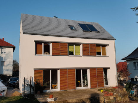 Ein modernes dreistöckiges Haus mit Schrägdach und Solaranlage, das an die eleganten Massivhäuser in Dresden erinnert. Die Fassade verfügt über große Fenster mit Holzläden und ist von einem kleinen Garten mit Steintreppen und Nachbarhäusern unter einem klaren blauen Himmel umgeben.