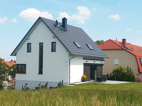 Ein modernes, weißes Haus mit einem dunkelgrauen, schrägen Dach steht auf einer Rasenfläche unter blauem Himmel. Dieses beeindruckende Beispiel eines Dresdner Massivhauses verfügt über vertikale Fenster und eine Terrasse mit Gartenmöbeln. Im Hintergrund sind andere Häuser zu sehen, die seinen Charme noch verstärken.