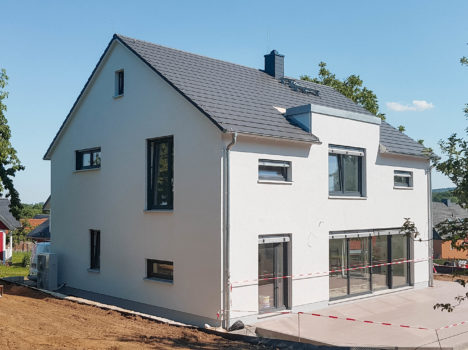 Ein modernes zweistöckiges Haus mit großen Fenstern und einem grauen Dach steht zwischen Bäumen und Nachbarhäusern und spiegelt den Charme des Dresdner Hausbaus wider. In der Nähe parkt ein Baufahrzeug, und der Bereich ist abgesperrt, was auf laufende Arbeiten hinweist. Der Himmel ist klar und sonnig.
