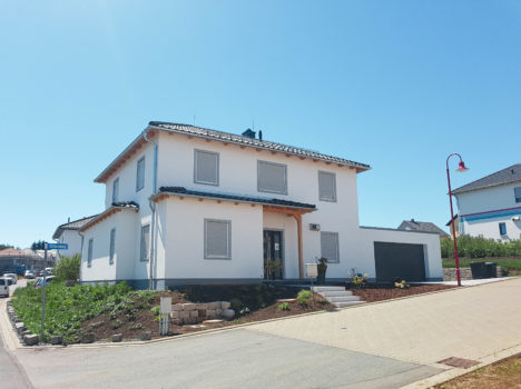 Auf einem Eckgrundstück in Sachsen steht ein modernes zweistöckiges weißes Haus mit grauem Dach. Das Massivhaus verfügt über geschlossene Fensterläden und eine Doppelgarage. Ein gepflegter Garten umgibt das Anwesen, während eine rote Straßenlaterne sich vom klaren blauen Himmel abhebt.
