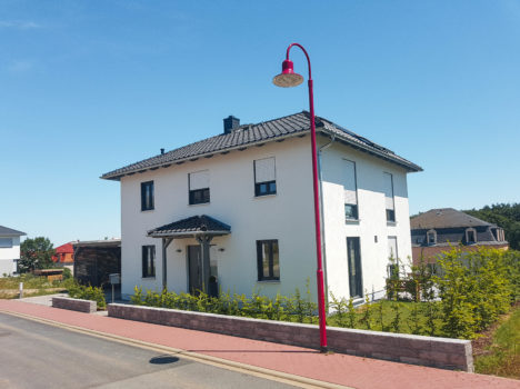 Ein zweistöckiges weißes Haus mit dunklem Dach und Fensterläden steht unter einem klaren blauen Himmel. Dieses charmante Massivhaus in Sachsen verfügt über einen kleinen Garten, der von einem niedrigen Zaun begrenzt wird. Am Rand des Gehwegs auf der linken Seite steht eine rote Straßenlaterne, in der Nähe befinden sich weitere Häuser.