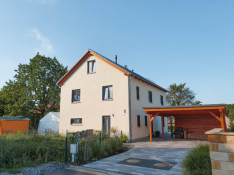 Ein modernes zweistöckiges Haus mit cremefarbenen Wänden und rotem Dach, das an ein Dresdner Massivhaus erinnert. Es verfügt über große Fenster, einen überdachten Carport auf der rechten Seite und ist von Grünflächen und einer gepflasterten Auffahrt umgeben. Der Himmel ist klar und blau und lässt auf einen sonnigen Tag schließen.