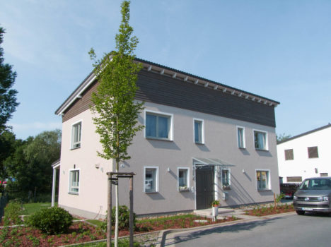 Dieses moderne zweistöckige Haus mit hellgrauen Wänden und dunklen Zierelementen ist ein beeindruckender Massivhausbau in Dresden. Er verfügt über große Fenster und einen glasüberdachten Eingang. Im Vorgarten steht ein junger Baum, umgeben von einem angelegten Garten. Auf der rechten Seite ist ein geparktes Auto zu sehen, das dem Haus noch mehr Charme verleiht.