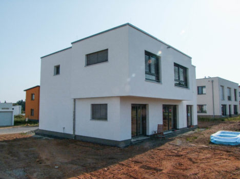 Auf einem unbefestigten Grundstück steht ein zweistöckiges, modernes weißes Haus mit großen Fenstern und einem Flachdach, das den Stil eines Dresdner Massivhauses verkörpert. In der Nähe des Eingangs stehen zwei kleine Stühle, während im sonnigen Hintergrund ähnliche Häuser und ein kleines aufblasbares Schwimmbecken zu sehen sind.