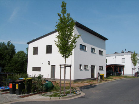 Ein modernes weißes zweistöckiges Haus mit Flachdach steht auf einem Eckgrundstück und zeigt den eleganten Stil der Massivhäuser Dresdens. Kleine Bäume und Sträucher schmücken den Hof, während an einem klaren Tag Recyclingbehälter und ein Müllcontainer am Straßenrand stehen.