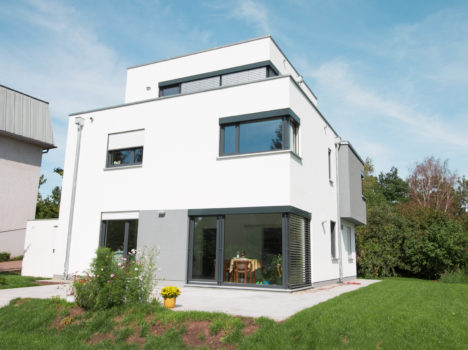 Dieses moderne, zweistöckige, weiße Massivhaus in Dresden verfügt über große Fenster und eine Gartenterrasse. Auf dem gepflegten Rasen steht neben den Glasschiebetüren ein Tisch mit Stühlen. Der klare, blaue Himmel unterstreicht das klare, zeitgenössische Design dieses eleganten Hauses in Sachsen.
