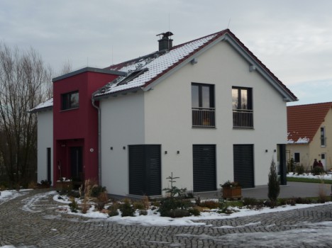 Ein modernes zweistöckiges Haus in Dresden mit einer weiß-roten Außenfassade, einem teilweise schneebedeckten Dach und dunklen Fensterläden. Dieser charmante Massivhausbau verfügt über einen kleinen Balkon und ist von einer gepflasterten Auffahrt und spärlicher Winterlandschaft umgeben – perfekt für alle, die elegantes Design zu schätzen wissen.