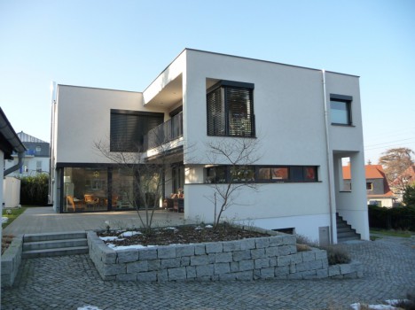 Ein modernes, zweistöckiges Dresdner Massivhaus mit Flachdach und großen Fenstern. Das Äußere ist minimalistisch und weiß. Dieses Massivhaus Sachsen verfügt über eine Terrasse mit minimaler Landschaftsgestaltung, einen Steinweg und Stufen, die zum Eingang führen. Die umliegenden Vorstadthäuser sind sichtbar.