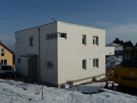 Ein modernes, weißes, würfelförmiges Haus steht majestätisch in der verschneiten Landschaft und verkörpert die Eleganz der Massivhäuser Dresdens. Ein in der Nähe geparktes Baufahrzeug deutet auf laufende Bauarbeiten hin. Umliegende Häuser und ein klarer blauer Himmel vervollständigen diese malerische Szene.