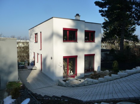 Modernes zweistöckiges kubisches Haus mit weißen Wänden und rot umrandeten Fenstern, das die Essenz eines Dresdner Massivhauses verkörpert. Es verfügt über eine gepflasterte Auffahrt und eine abgestufte Steinlandschaft. Im Hintergrund sind unter einem klaren blauen Himmel hohe Nadelbäume zu sehen.