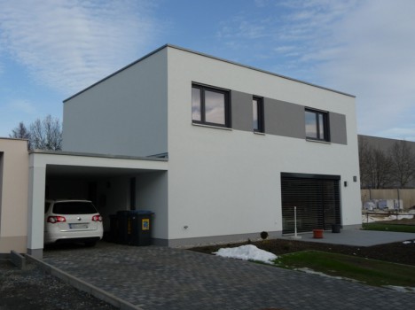 Modernes zweistöckiges Haus mit Flachdach, sauberer weißer Fassade und minimalistischem Design, das den anspruchsvollen Stil des Massivhausbaus Dresden verkörpert. Ein Auto ist unter dem angeschlossenen Carport geparkt und die Auffahrt ist mit Ziegeln gepflastert. Schneeflecken auf dem Boden lassen eine ruhige Winterlandschaft erahnen.