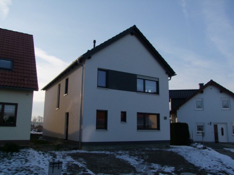 Ein zweistöckiges Haus mit dunkelgrauem Dach und modernem Design mit großen Fenstern, typisch für den Hausbau in Dresden, liegt zwischen zwei ähnlichen Häusern. Der Boden ist teilweise mit Schnee bedeckt, während der klare Himmel seinen zeitgenössischen Charme verstärkt.