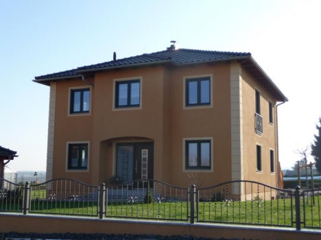 Ein zweistöckiges Haus mit beigen Wänden und einem dunklen Ziegeldach, mit mehreren Fenstern und einer Eingangstür mit Glaseinsätzen, spiegelt den anspruchsvollen Stil des Massivhausbaus Dresden wider. Es ist von einem grünen Rasen umgeben und von einem schwarzen Metallzaun umschlossen.
