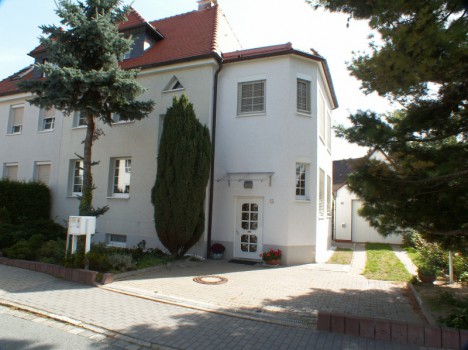 Ein weißes, zweistöckiges Dresdner Massivhaus mit einem Dachgeschoss mit Schrägdach und großen Fenstern. Davor steht ein großer immergrüner Baum, neben Sträuchern und einem gepflegten Rasen. Eine Auffahrt führt zu einer kleinen Garage auf der rechten Seite und zeigt den Charme des Massivhaus Sachsen-Designs.