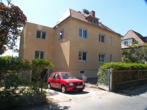 Hinter einer kleinen Einfahrt steht ein zweistöckiges beiges Haus mit rotem Dach. Davor parkt ein rotes Auto, das den Stil der Massivhäuser Dresden widerspiegelt. Das von Grün und einem Metallzaun umgebene Anwesen profitiert von den nahe gelegenen Bäumen, die an sonnigen Tagen Schatten spenden.