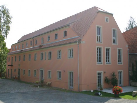 Ein großes, rechteckiges, pfirsichfarbenes Gebäude mit mehreren Fenstern steht vor einem klaren Himmel. Dieses Beispiel für Massivhausbau in Dresden verfügt über ein rotes Ziegeldach und Pflanzen rund um den Eingang. Im Vordergrund vervollständigen ein Kopfsteinpflaster und eine schwarz-weiße Katze diese bezaubernde Szene.