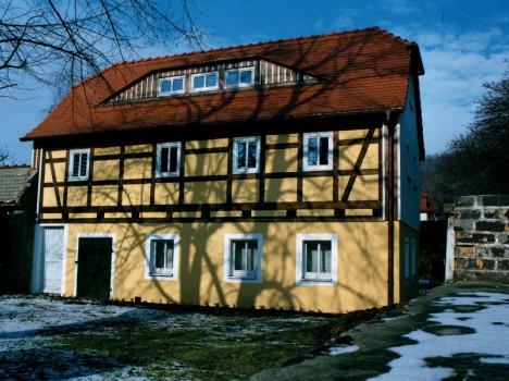 Dieses charmante mehrstöckige Massivhaus in Sachsen besticht durch ein rotes Ziegeldach und gelbe Wände mit braunen Fachwerkbalken. Unter einem klaren blauen Himmel werfen kahle Äste Schatten auf die Fassade, während Schneeflecken den Boden bedecken.