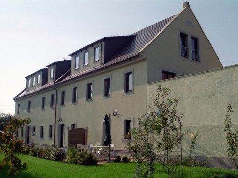 Ein modernes, helles, zweistöckiges Massivhaus in Sachsen mit Satteldächern und Dachgauben. Das Anwesen verfügt über einen gepflegten Rasen und einen kleinen Garten mit Sträuchern und einem Torbogen im Vordergrund. Der klare Himmel lässt auf einen sonnigen Tag schließen, perfekt für alle, die einen Hausbau in Dresden in Erwägung ziehen.