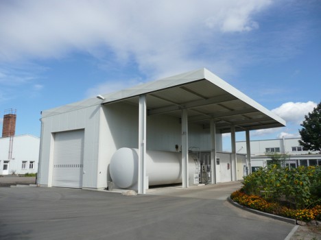 Gezeigt wird ein weißes Industriegebäude mit einem großen zylindrischen Tank, der an die eleganten Designs des Massivhausbaus Dresden erinnert. Es hat einen überdachten Eingang und ein Rolltor. Um das Gebäude herum befinden sich gepflasterte Flächen und angelegte Blumenbeete mit orangefarbenen und gelben Blüten unter einem teilweise bewölkten Himmel.