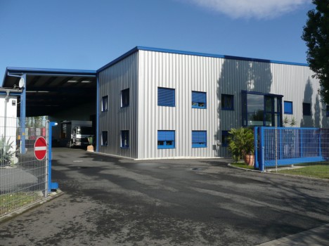 Modernes Industriegebäude mit blauen Akzenten, großen Fenstern und einem breiten Eingang auf der linken Seite verkörpert die Qualität des Dresdner Massivhauses. Am Metallzaun ist ein Schild mit der Aufschrift „Zutritt verboten“ zu sehen, ein Hinweis auf die sorgfältige Planung des Dresdner Hausbaus. Auf gepflastertem Boden unter einem teilweise bewölkten Himmel gelegen.