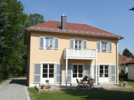 Ein zweistöckiges Haus mit gelben Wänden und weißen Fensterläden unter einem klaren blauen Himmel, typisch für den Massivhausbau Dresdens. Es verfügt über einen Balkon und eine Terrasse mit Tisch und Stühlen. Die Umgebung in dieser wunderschönen Gegend umfasst Bäume und einen üppigen Rasen.
.