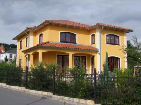 Dieses charmante zweistöckige gelbe Haus, ein Dresdner Massivhaus, verfügt über ein rotes Ziegeldach und gewölbte Fenster, anmutig umgeben von einem Metallzaun und üppigem Grün. Der bewölkte Himmel verleiht der Szene Tiefe, und dahinter ist ein weiteres Haus zu sehen.