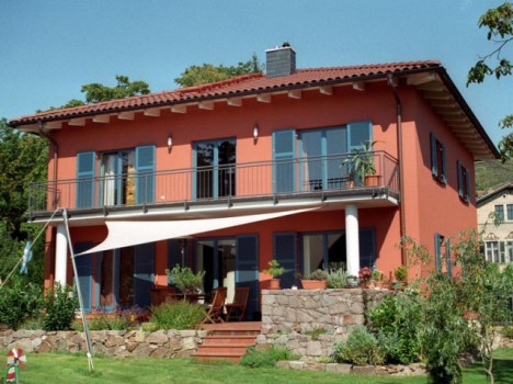 Ein zweistöckiges Haus mit roter Außenfassade und Ziegeldach sowie blauen Fensterläden. Es verfügt über einen Balkon und eine Terrasse mit Sonnenschirm. Dieses charmante Fertighaus Dresden ist von einem gepflegten Garten und einer Steinmauer umgeben und steht unter klarem Himmel.