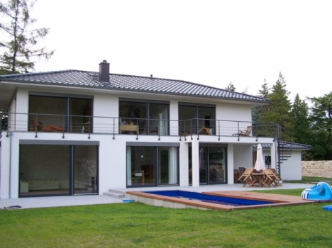 Ein modernes, zweistöckiges Massivhaus mit großen Glastüren und einem Balkon mit Blick auf einen rechteckigen Pool im Hinterhof. Das in Dresden gelegene Haus ist von Gras und Bäumen umgeben und verfügt über einen Terrassenbereich mit Gartenmöbeln, der die Eleganz der sächsischen Architektur verkörpert.