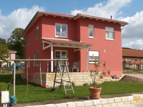Ein charmantes zweistöckiges rotes Haus mit einer kleinen Veranda steht inmitten eines üppigen grünen Rasens und erinnert an ein klassisches Dresdner Massivhaus. Eine Topfpflanze schmückt den Vordergrund, während eine Schaukel zum Spielen einlädt. Der Himmel ist teilweise bewölkt, was die gemütliche Atmosphäre dieses modernen Rückzugsorts noch verstärkt.