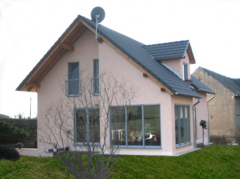 Ein modernes zweistöckiges Haus mit beiger Fassade und grauem Satteldach, das einem typischen Dresdner Massivhaus ähnelt. Es verfügt über große Fenster auf beiden Etagen und einen Balkon. Im Vordergrund steht ein blattloser Baum, ergänzt durch eine Satellitenschüssel auf dem Dach.
