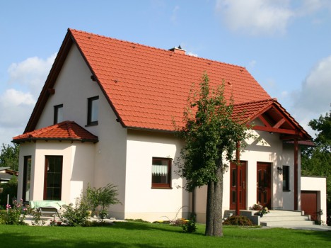 Ein kleines, weißes Haus mit rotem Satteldach ragt stolz vor einem blauen Himmel mit flauschigen Wolken empor. Dieses charmante Massivhaus in Sachsen ist von einem gepflegten Rasen und einigen Bäumen umgeben und schafft eine friedliche, vorstädtische Szenerie.