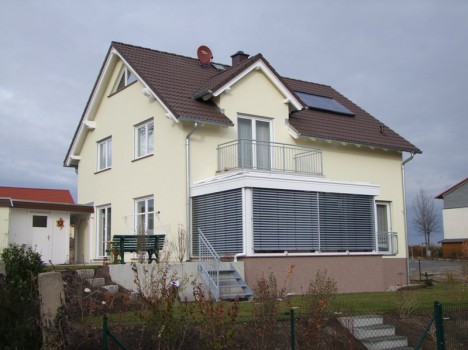 Ein zweistöckiges beiges Haus mit einem braunen Dach verfügt im unteren Stockwerk über einen mit Jalousien abgeschirmten Wintergarten. Auf dem Dach ist eine Satellitenschüssel montiert. Das im Stil des Dresdner Massivhauses erbaute Haus führt über eine kleine Treppe in den Garten mit einer Bank und blattlosen Sträuchern im Vordergrund.