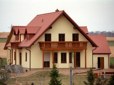 Ein zweistöckiges Haus mit cremefarbenen Wänden und einem roten Ziegeldach, erbaut im Stil der Dresdner Massivhäuser. Es verfügt über einen Balkon, mehrere Fenster und eine Veranda. Das Haus liegt eingebettet in einer ländlichen Gegend mit Feldern und vereinzelten Bäumen im Hintergrund.