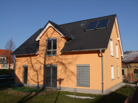 Dieses zweistöckige, pfirsichfarbene Dresdner Massivhaus mit seinem dunklen Dach, auf dem sich Solarzellen befinden, verfügt über mehrere Fenster mit Fensterläden. Ein großer Baum wirft einen Schatten auf die Vorderseite und unter einem klaren blauen Himmel sind Rasen und Dach mit Schneeflecken bedeckt.