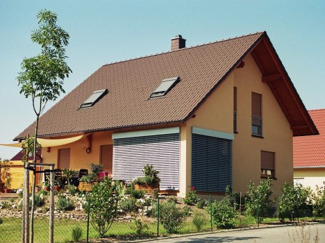 Ein zweistöckiges Haus mit dunkelbraunem Satteldach, cremefarbenen Wänden und großen Fenstern mit Fensterläden verkörpert den eleganten Stil eines sächsischen Massivhauses. Der Vorgarten voller verschiedener Pflanzen und Blumen ergänzt den klaren blauen Himmel und den stolz im Vordergrund stehenden Baum.