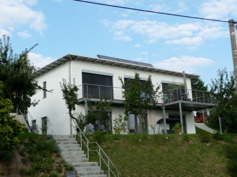 Ein modernes zweistöckiges weißes Haus in Dresden verfügt über Solarmodule auf dem Dach, große Fenster mit Fensterläden und einen Balkon. Dieser Massivhausbau ist von üppigem Grün umgeben und über eine Treppe an einem Grashang zugänglich. Der Himmel ist teilweise bewölkt, was die ruhige Lage noch verstärkt.