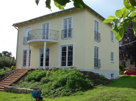 Ein zweistöckiges gelbes Dresdner Massivhaus mit grünem Dach und geschwungenem Balkon steht auf einer Rasenfläche. Es verfügt über große weiß gerahmte Fenster und eine Holztreppe, die zum Eingang führt und seinen architektonischen Charme noch verstärkt. Ein rotes Auto parkt an der Seite und vervollständigt diese malerische Szene.