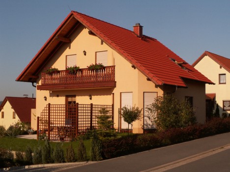 Ein beiges zweistöckiges Haus mit rotem Satteldach, hölzernem Balkongeländer und einem Spalier an der Seite verkörpert den Charme des Dresdner Massivhauses. Eingebettet zwischen kleinen Bäumen und Sträuchern im Vorgarten steht es anmutig an einer abschüssigen Straße unter einem klaren blauen Himmel.