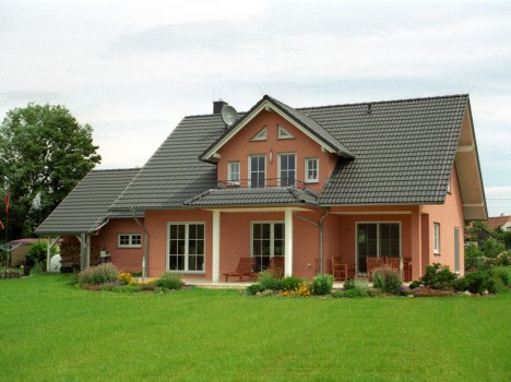 Ein großes, pfirsichfarbenes Haus mit einem dunkelgrauen Ziegeldach steht auf einem üppigen grünen Rasen und verkörpert den Charme eines Dresdner Massivhauses. Das Gebäude verfügt über mehrere Fenster und einen kleinen Balkon. In der Nähe des Eingangs sind unter einem bewölkten Himmel Sträucher und Blumen gepflanzt.