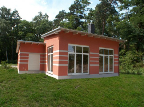 Ein modernes, einstöckiges Haus mit Flachdach und großen Fenstern, in warmem Korallenrot mit weißen Streifen gestrichen. Dieses Dresdner Massivhaus steht auf einer Rasenfläche, umgeben von hohen Bäumen unter einem klaren Himmel.