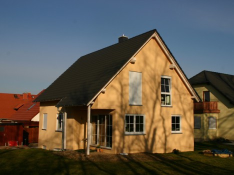Ein hellgelbes zweistöckiges Haus mit einem dunklen Schrägdach steht inmitten eines grasbewachsenen Hofs und verfügt über Fenster auf beiden Etagen. Unter dem klaren blauen Himmel und den Schatten der Bäume, die auf seine Wände fallen, verkörpert es die Eleganz des Massivhausbaus in Dresden.