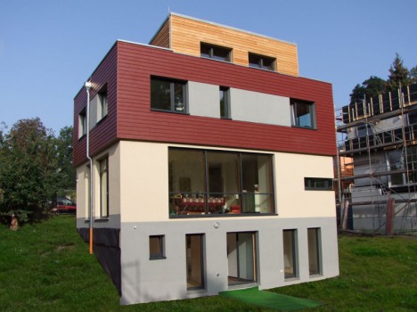 Ein modernes, mehrstöckiges Haus mit Flachdach und einer Mischung aus Beige-, Braun- und Holzverkleidung ragt stolz zwischen den Massivhäusern Dresden hervor. Große Fenster auf jeder Etage bieten Ausblicke auf eine Rasenfläche. In der Nähe deutet die Entwicklung eines teilweise fertiggestellten Gebäudes auf das Wachstum von Fertighäusern Dresden hin.