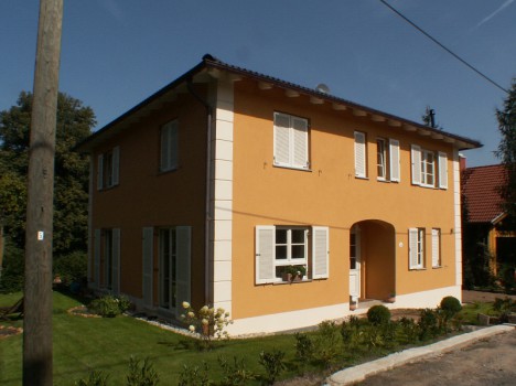 Ein zweistöckiges Haus mit orangefarbener Fassade und weißen Fensterläden, eingebettet in einen üppigen grünen Rasen mit Sträuchern. Der Vordereingang verfügt über einen Torbogen vor einem klaren blauen Himmel. Dieses charmante Wohnhaus fängt die Essenz des Hausbaus in Dresden ein.
