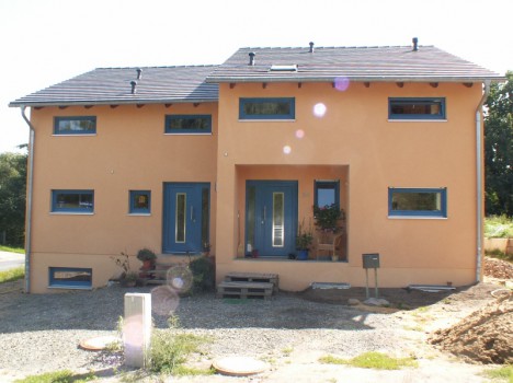 Ein zweistöckiges, pfirsichfarbenes Dresdner Massivhaus verfügt über ein dunkelgraues Dach, blau gerahmte Fenster und eine blaue Eingangstür. Topfpflanzen schmücken den Eingang, und ein Kiesweg führt dorthin. Sonnenlicht erzeugt auf dem Bild Lichtreflexe, die seinen ruhigen Charme verstärken.