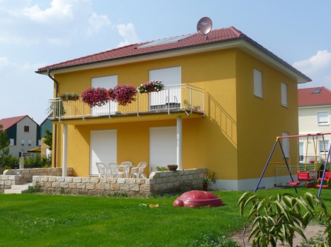 Ein charmantes, zweistöckiges gelbes Haus mit rotem Dach und Balkon, das mit leuchtend rosa und roten Blumen geschmückt ist. Davor befindet sich eine gemütliche Terrasse mit weißen Stühlen und einem Tisch. In der Nähe, auf dem Rasen, vergnügen sich die Kinder auf einer Schaukel – ein perfektes Beispiel für den beliebten Dresdner Massivhausbaustil.