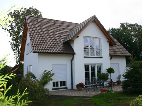 Ein modernes zweistöckiges Haus in Dresden mit einem schrägen braunen Ziegeldach und einer weißen Außenfassade besticht durch große Fenster und eine mit Topfpflanzen geschmückte Terrasse. Dieses Beispiel eines Dresdner Massivhauses ist von üppigem Grün unter einem teilweise bewölkten Himmel umgeben und eignet sich perfekt zum Entspannen im Freien am Tisch mit Stühlen.