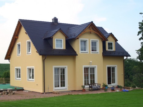Ein zweistöckiges gelbes Haus, das einem Dresdner Massivhaus ähnelt, mit einem dunkelgrauen Satteldach und mehreren Dachgauben. Das Haus verfügt über eine Terrasse mit Gartenmöbeln und ist von einem grünen Rasen und Bäumen im Hintergrund umgeben.