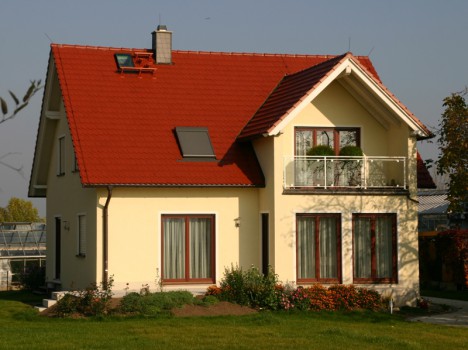 Dieses FertigHaus Dresden ist ein charmantes zweistöckiges Haus mit cremefarbenen Wänden und einem roten Satteldach. Es verfügt über große Fenster, einen gemütlichen, mit Pflanzen geschmückten Balkon und einen gepflegten Garten voller Blumen und Sträucher unter dem klaren Himmel.