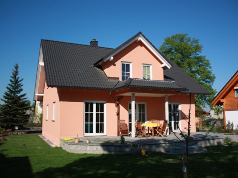Ein pfirsichfarbenes, zweistöckiges Haus mit dunklem Dach, typisch für den eleganten Dresdner Massivhausstil, verfügt über eine überdachte Terrasse mit Gartenmöbeln. Es ist von einem grünen Rasen umgeben, im Hintergrund sind unter einem klaren blauen Himmel ein Baum und ein weiteres Haus zu sehen.