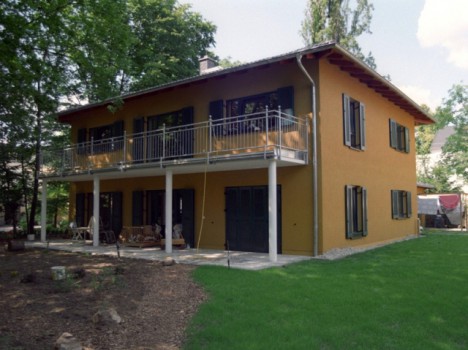 Dieses zweistöckige gelbe Haus, das an die eleganten Massivhäuser Dresdens erinnert, ist von üppigem grünem Gras und Bäumen umgeben. Die obere Etage verfügt über dunkle Fensterläden und einen von Säulen getragenen Balkon. Das Erdgeschoss bietet einen gemütlichen Terrassenbereich mit Gartenmöbeln, perfekt zum Genießen der Natur.